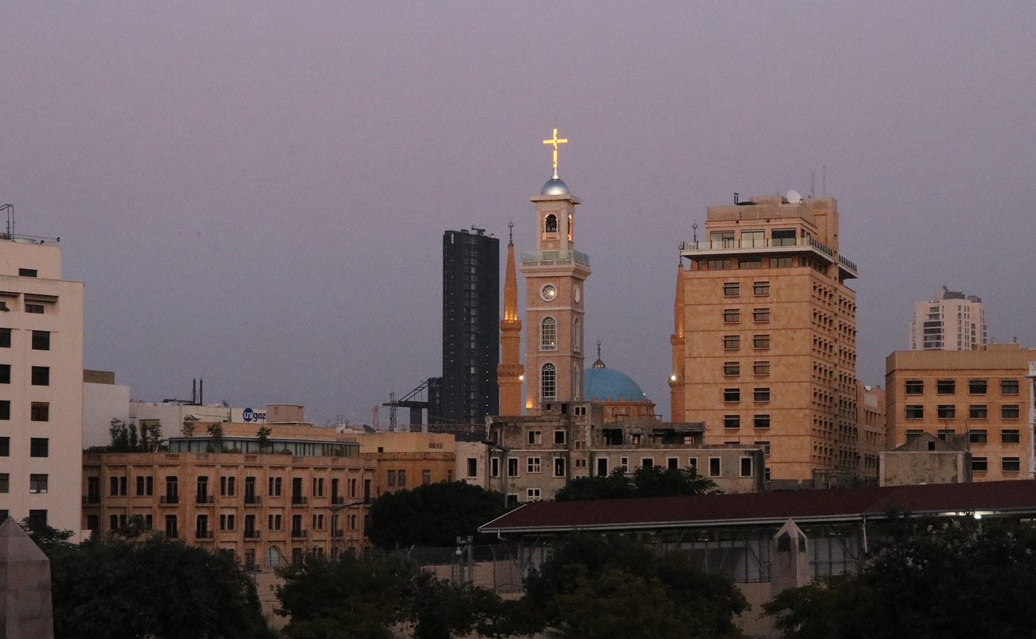 An illuminated cross sits atop a tall church spire, surrounded by apartment and office buildings.