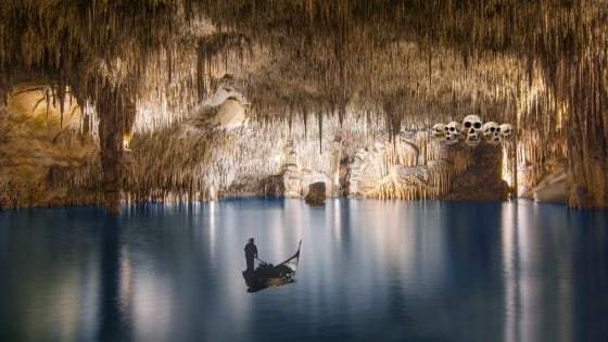 The ferryman poles his empty barge across an immense, glassy lake toward a cavernous shoreline a-drip with stalactites and a myriad straw-like speleothems. Skulls loom over the cave entrance at the edge of the water.