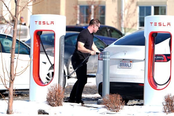 A man wearing a T-shirt plugs in a white car at a charging station.