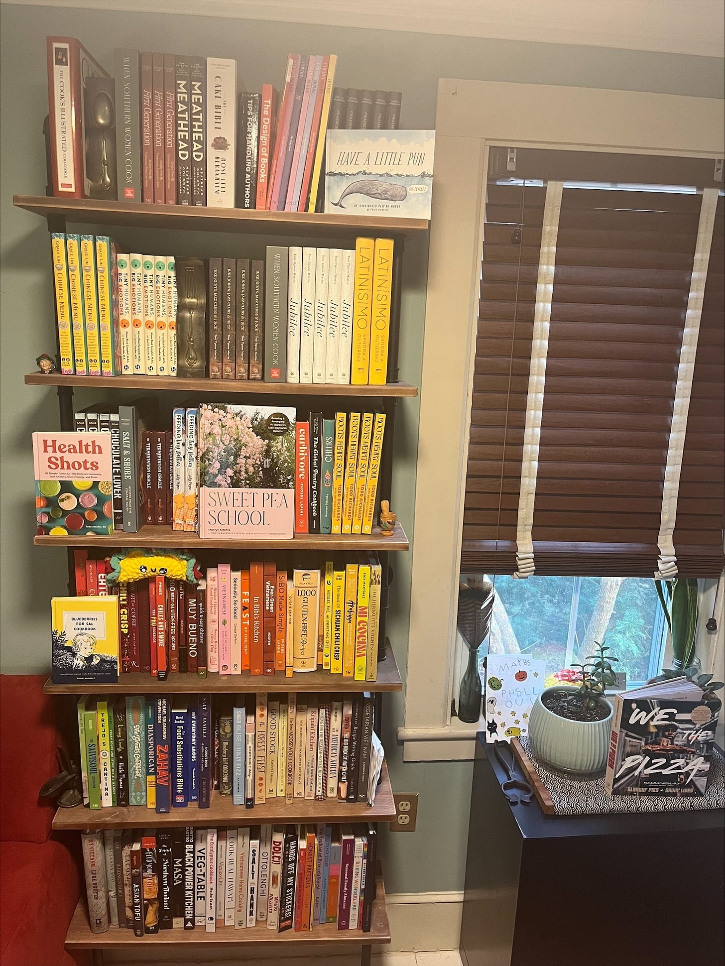 Cookbooks on wooden shelving next to a window with closed blinds.