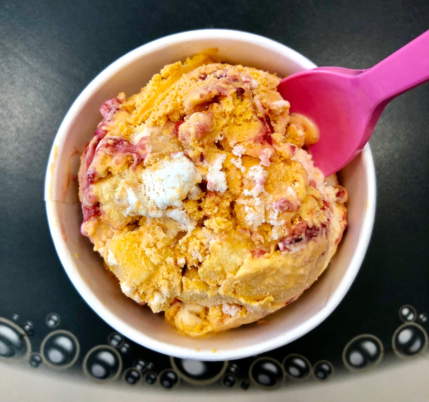 a scoop of orange and white ice cream with a berry-red swirl, in a white cup, with a pink spoon