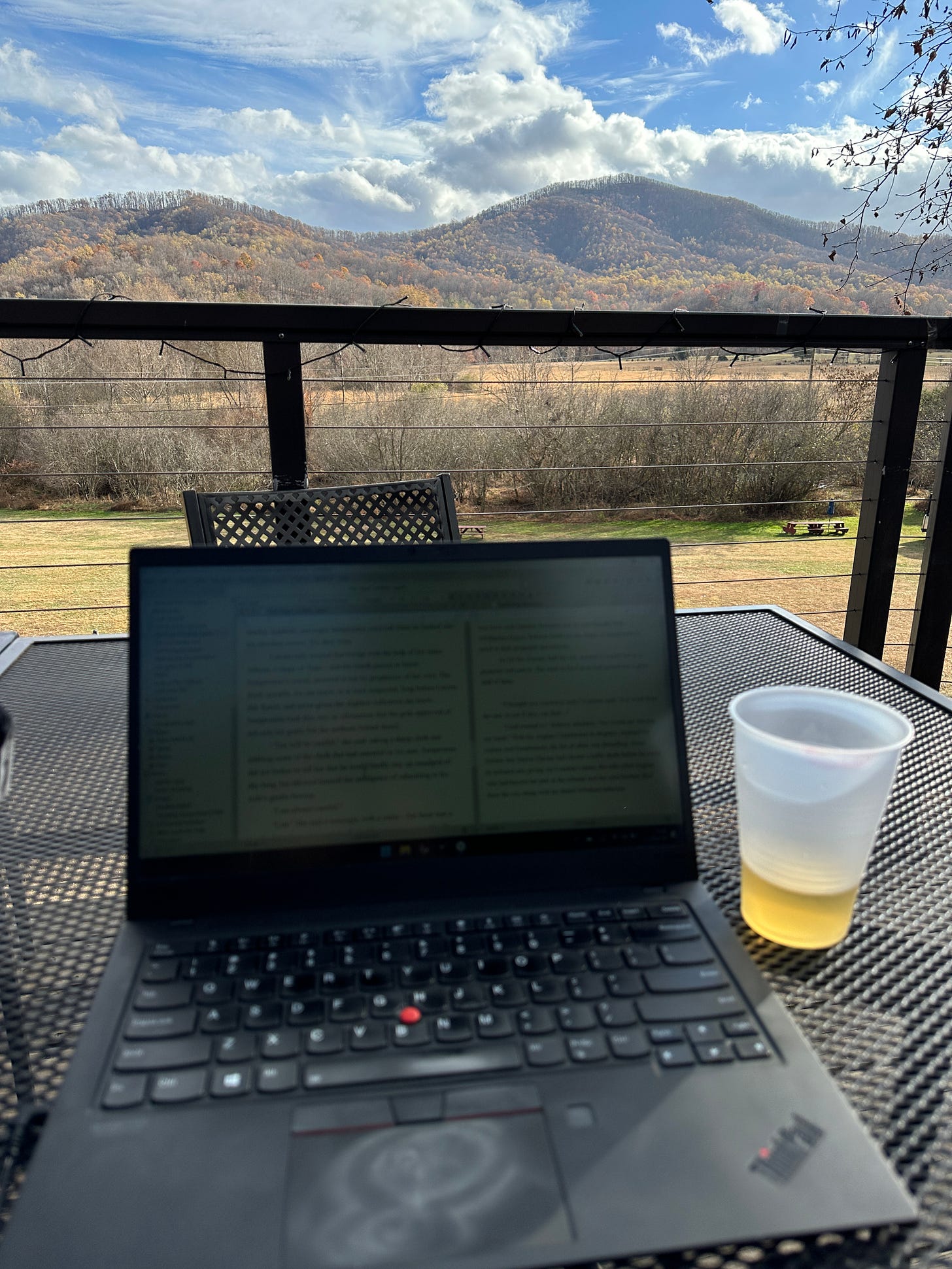 A laptop and a cup of cider on an iron-wrought table, a gorgoeous view of mountains and a cloud-streaked sky in the background