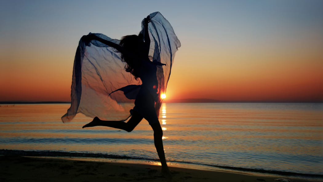 A dancer leaps along the sunset-kissed ocean shore, trailing a sheer veil like wings.
