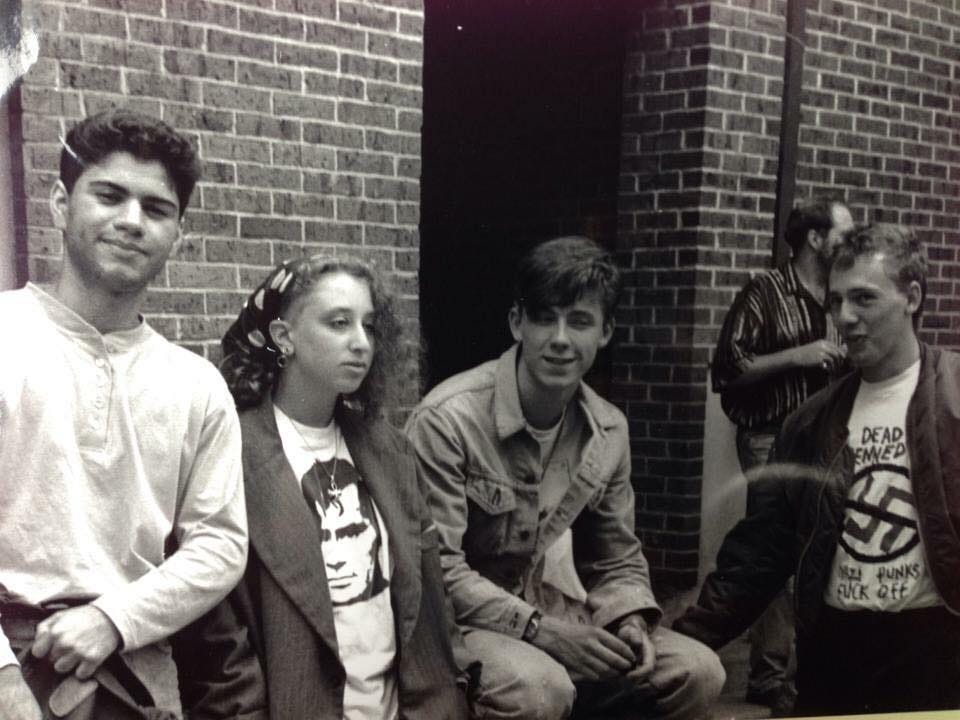 Three teenage boys and a teenage girl in front of a brick wall. Black and white photo. The girl wears a gray blazer over a white shirt with Jack Kerouac's image on the front