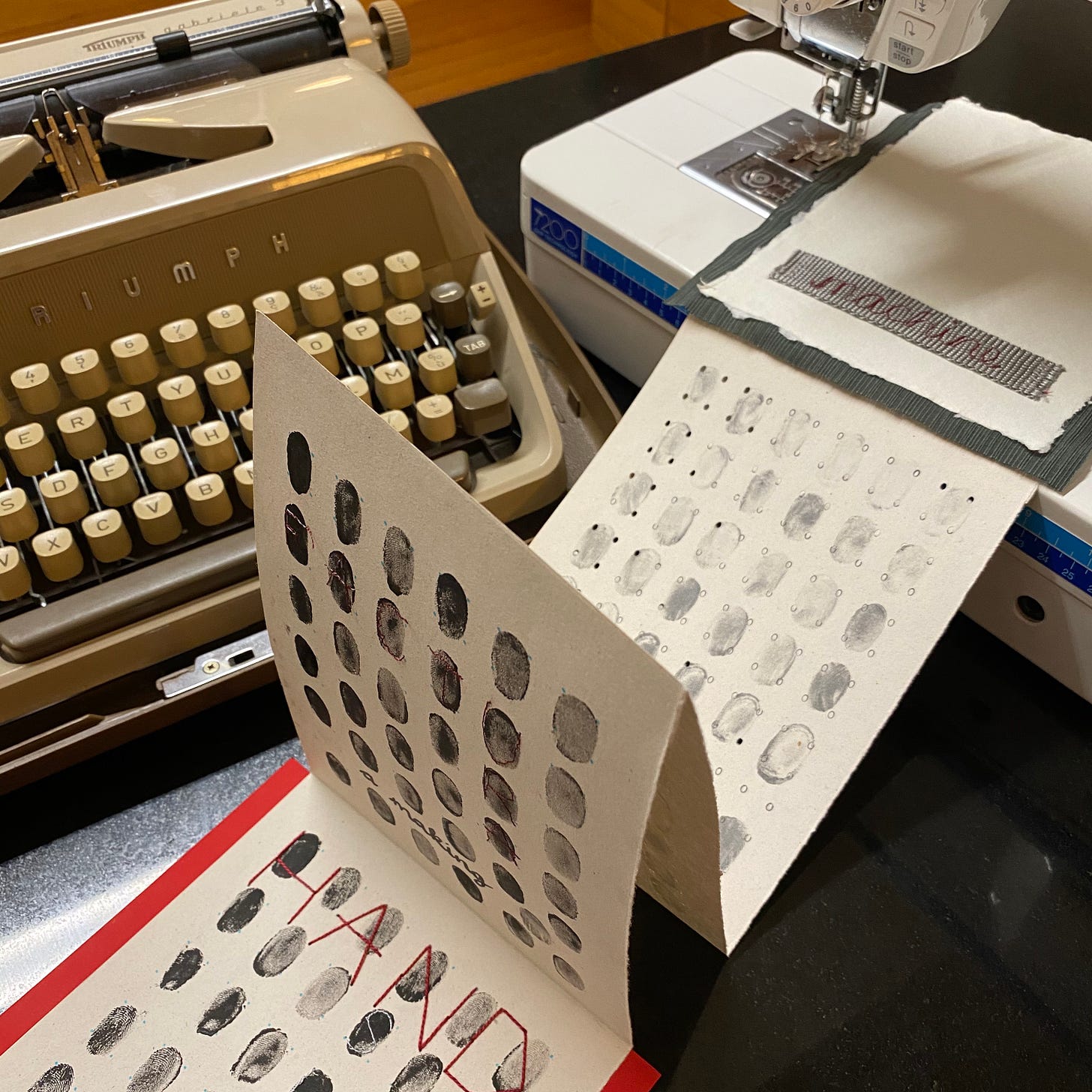 a handmade concertina book titled machine with thumbprints and other regular patterns on the pages created by typewritten letters repeated and stitched on a sewing machine - both the typewriter and the sewing machine are in the image.