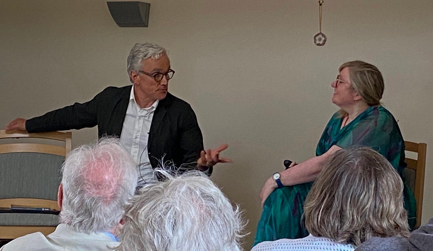 A white man and a white woman in conversation in front of an audience. A white man and a white woman in conversation in front of an audience.