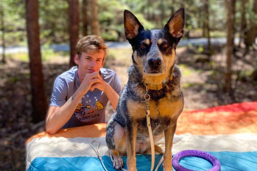 Scout the blue heeler poses atop a rainbow-colored Rumpl blanket draped across a campground picnic table. In the background, slightly blurred, Sean sits at the table bench and looks at the camera too