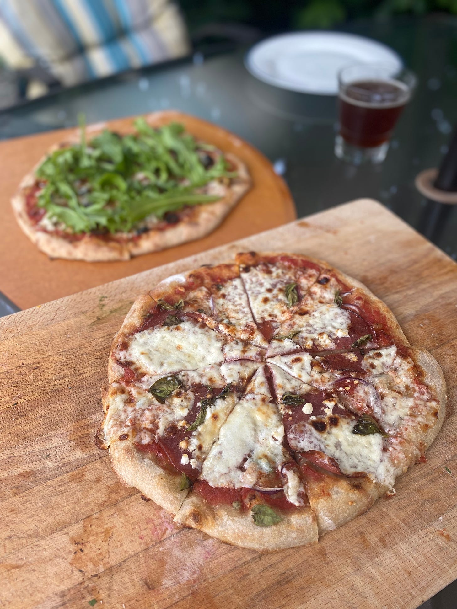Two pizzas on boards on an outdoor table. The nearest one has bubbled spots of mozza, basil leaves, and salami over a red sauce. The one in the background is blurred and scattered with arugula.