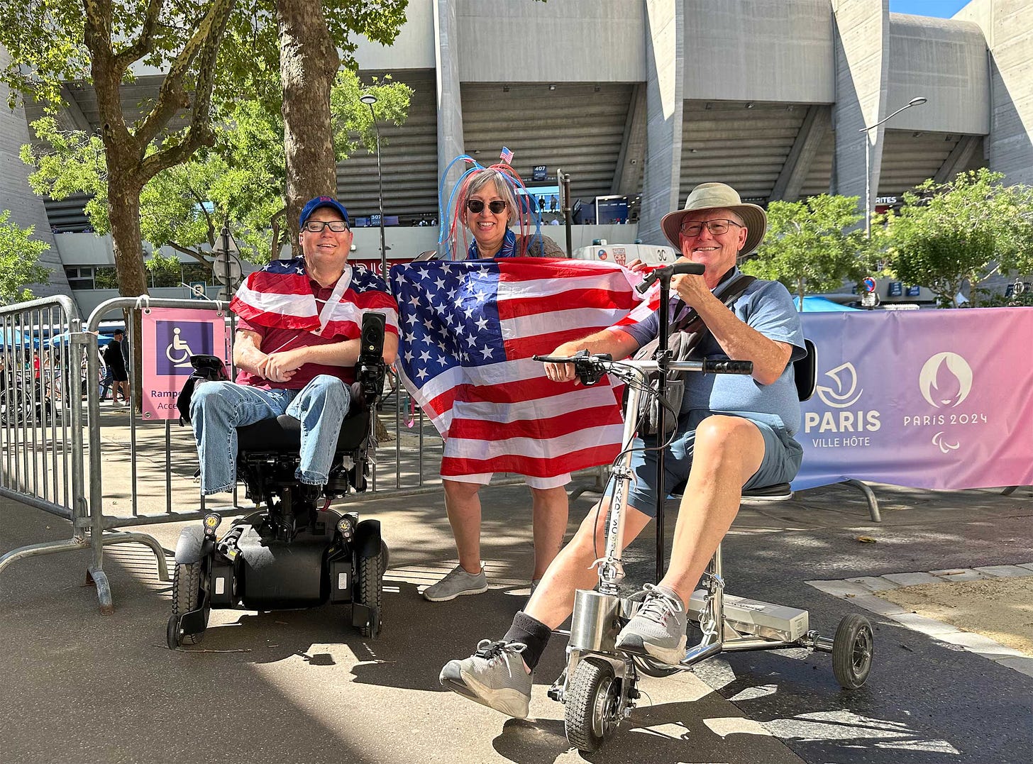 John seated in his wheelchair next to a man with a scooter and a woman holding a United States flag outside of a football stadium.