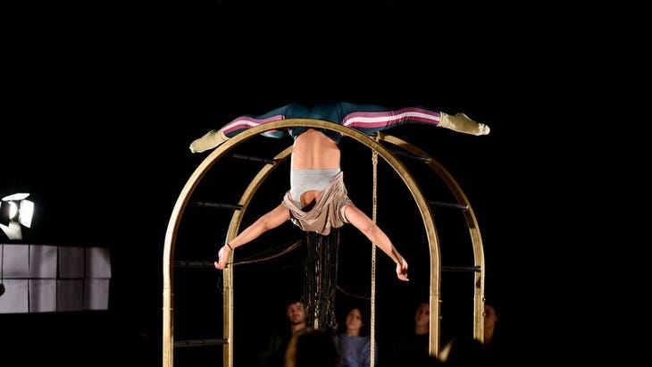 A performer hands upside down while holding on with their legs on a gold arched frame.