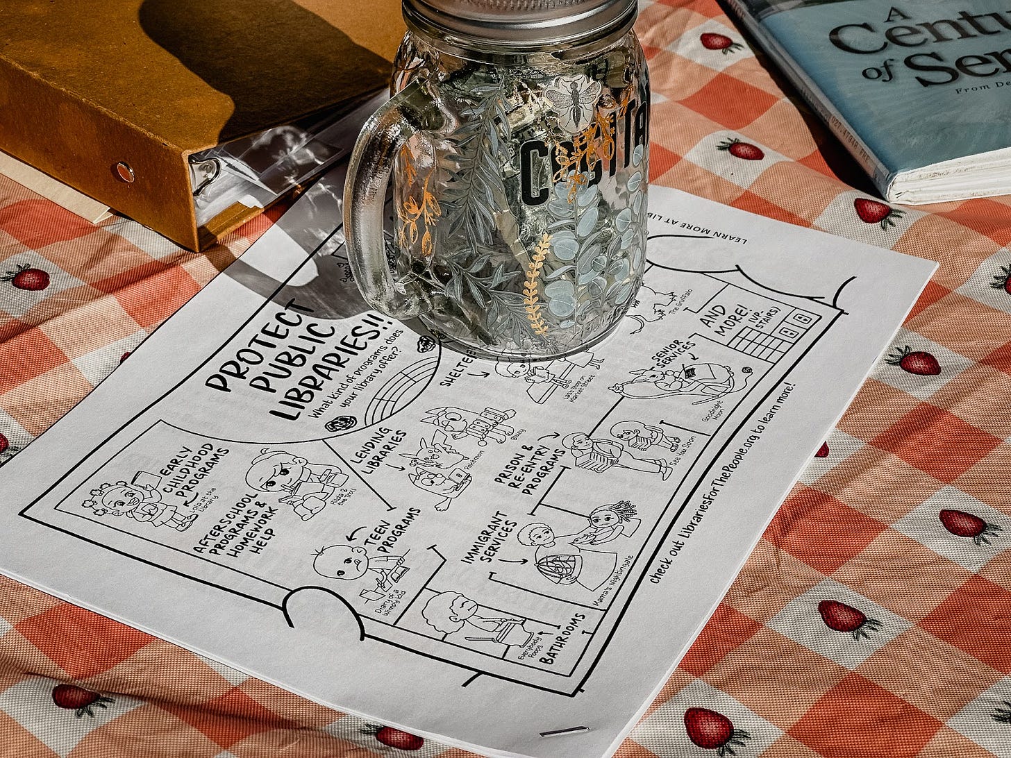 A close up photo of a lemonade stand table with an orange and white gingham/checkered tablecloth with strawberries in the white spots. There is a brown binder in the corner as well as a book. There is a mason jar with a handle on top of a printout about why protecting public libraries is important that is illustrated in a cartoon style