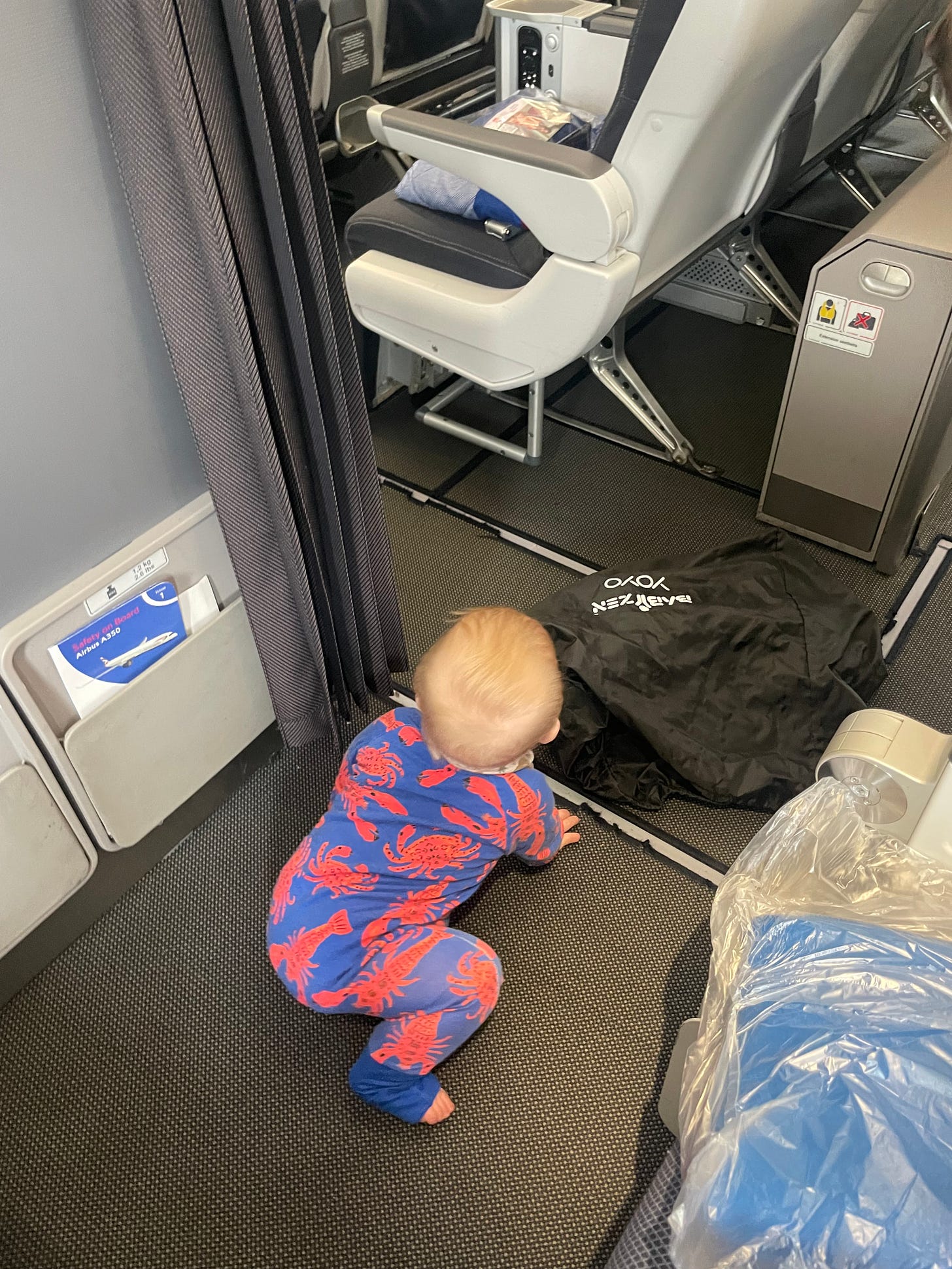 A baby in a blue and red lobster onesie sits on the floor of the plane, looking away from the camera, about to crawl off.