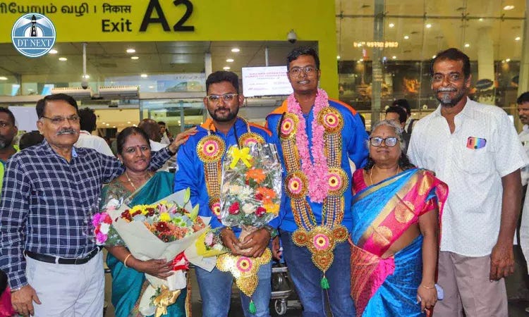 Photo of Sai Akash, Vice Caption of the Deaf T20 Indian Cricket Team, and E Sudarsan, member of the team as they are received by their families outside the Chennai Airport. Sai Akash & Sudarsan are standing in their jerseys flanked by their parents, to the left and right. Both parents are wearing garlands, and Sai Akash and hi mother are holding bouquets of flowers in their hands. 