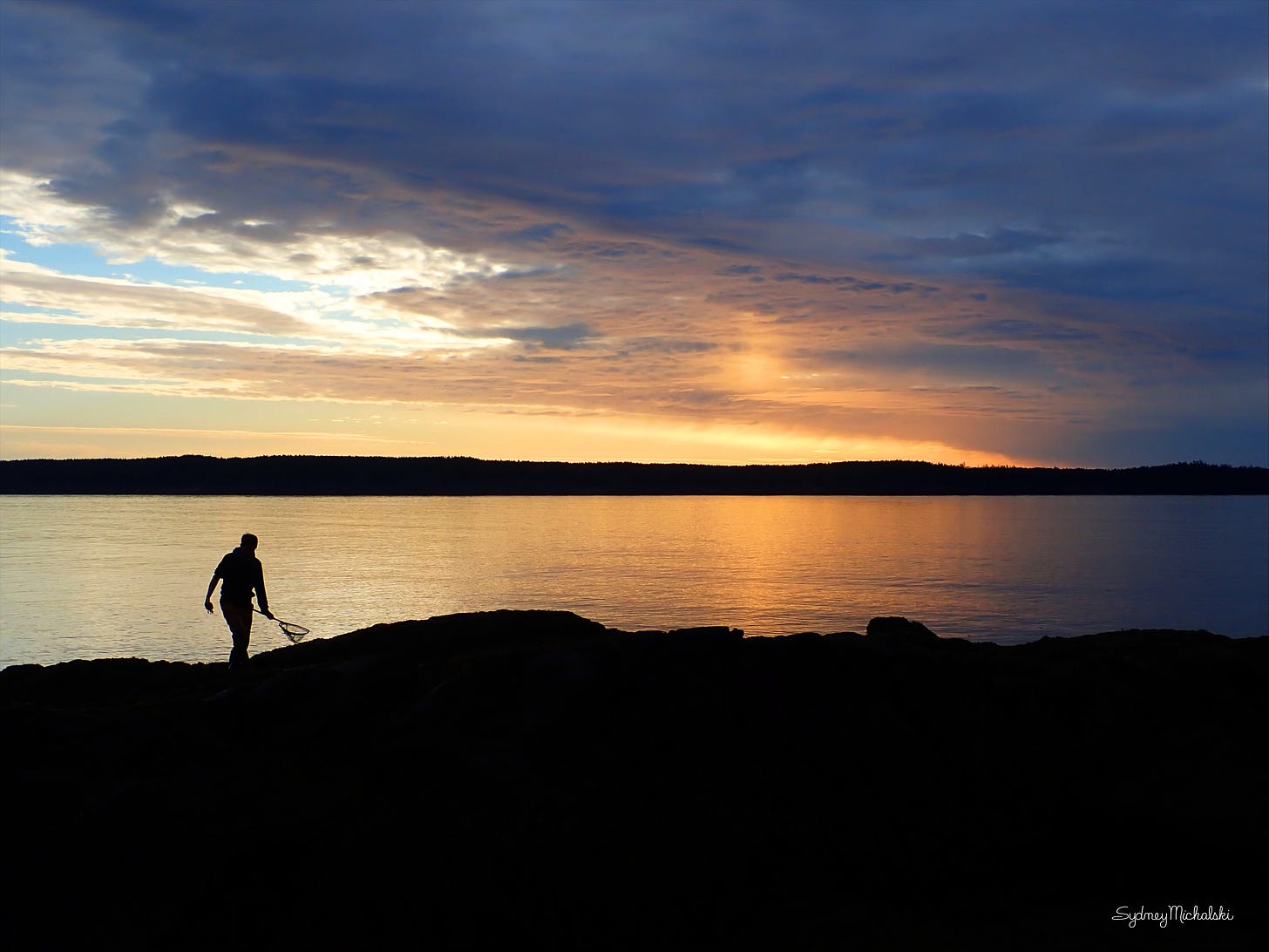 The silhouette of a young man, fishing net in hand, explores a rocky coastline at sunrise.