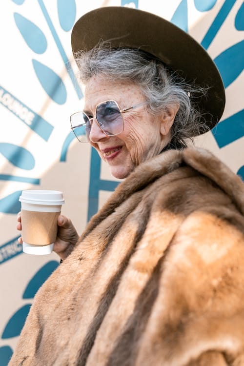 Free Elegant senior woman enjoying coffee outdoors, wearing a fur coat and hat, smiling. Stock Photo