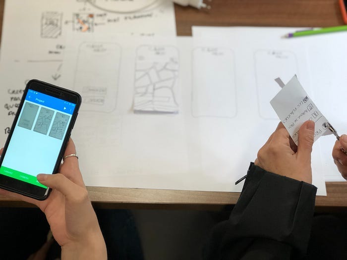Two hands holding different things. The hand on the left is holding a phone with a prototype loaded, with screenshots of paper. The hand on the right is cutting paper ‘screens’ to fit. The table has a whole bunch of paper sketches inside phone frames.