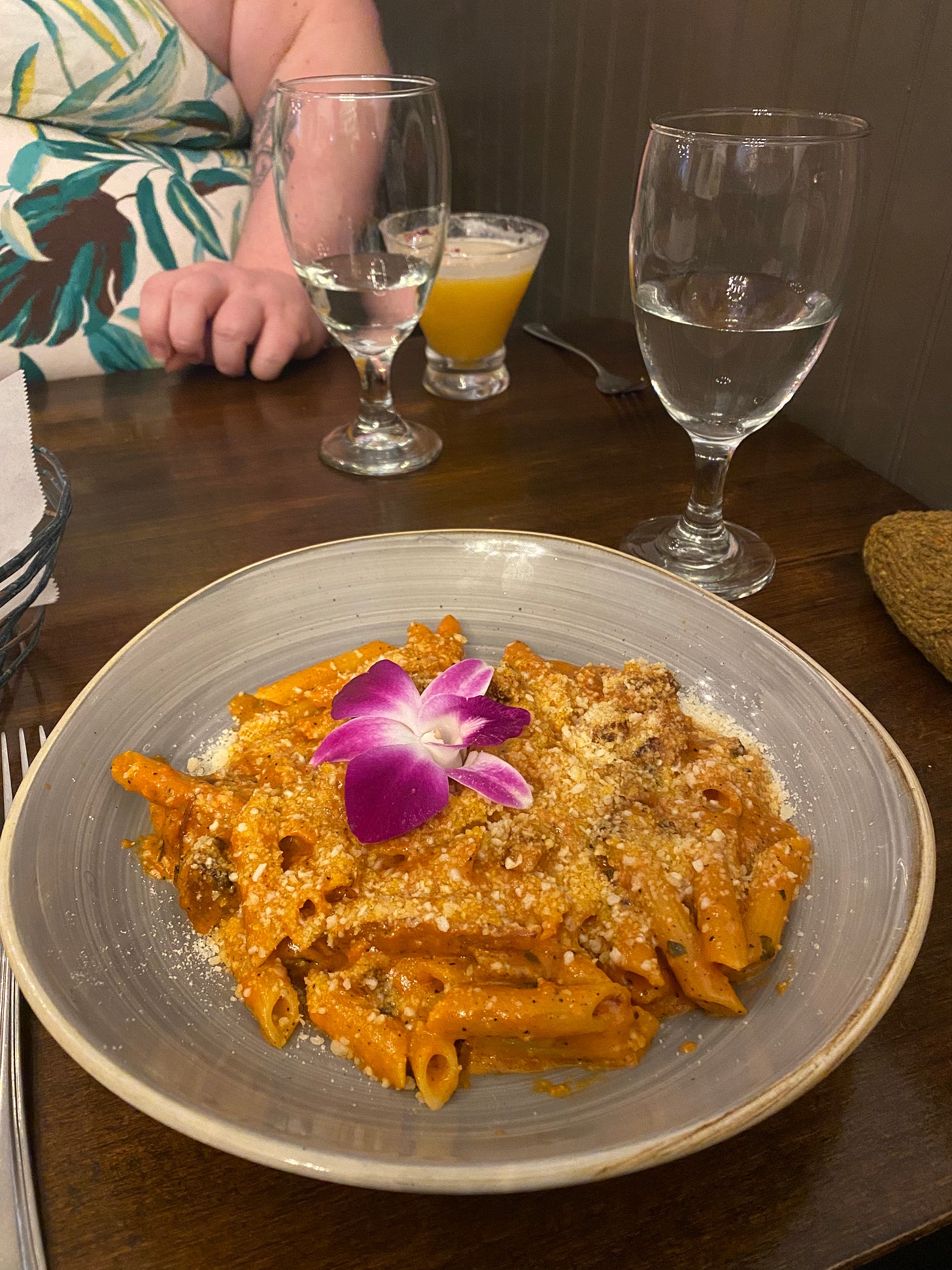 The penne alla vodka described above, in a blue-grey ceramic bowl with parmesan and a purple edible flower on top. Claire is just visible across the table, wearing a blue and white leaf print dress.