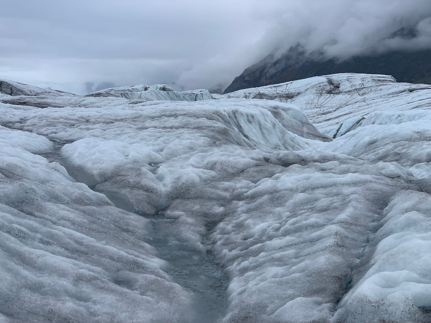 A photo of a glacier taken low to the ground so you see the glacier spread to meet a cloudy sky of similar hues