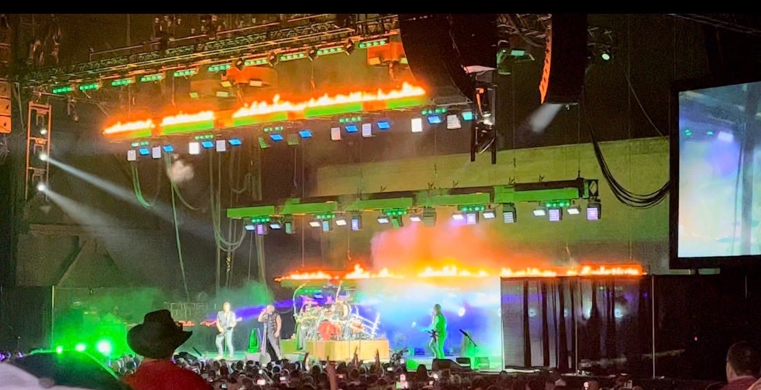 Disturbed performs near Portland, OR. Emerald and cobalt blue lights shine behind the band with flames burning behind them.