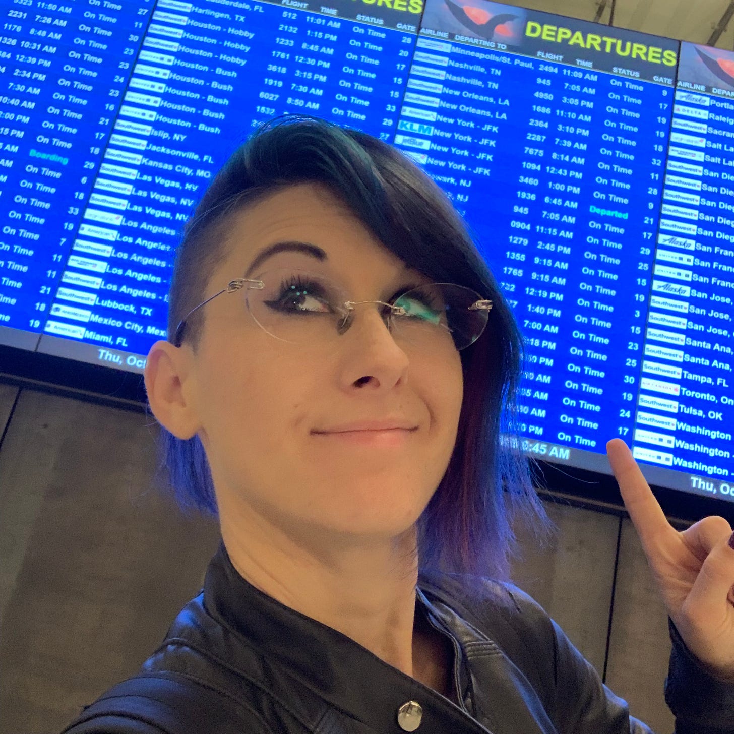 Lyric in an airport, pointing at the blue board of flights behind them.