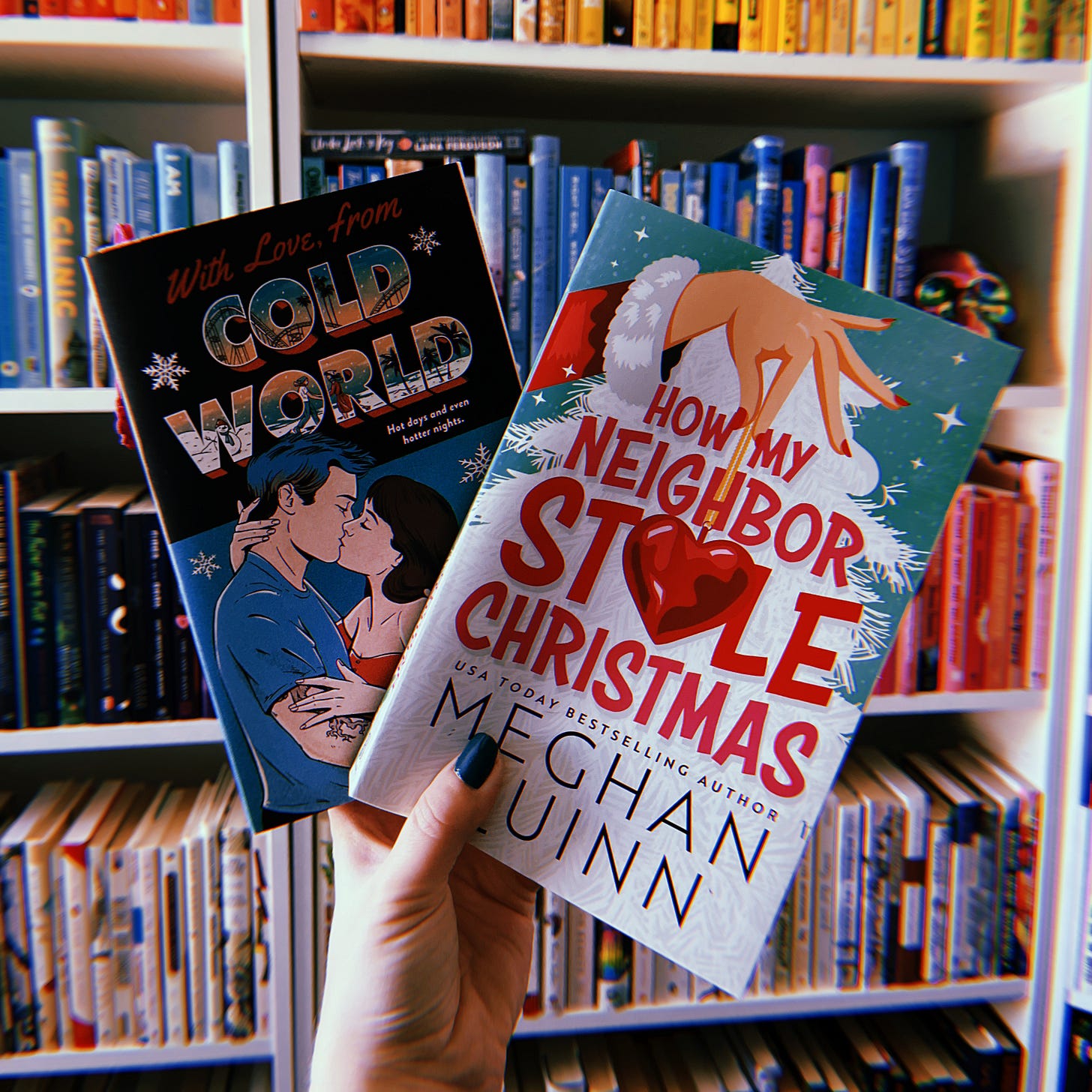 A hand holding up two books in front of a book shelf. The books are With Love, From Cold World and How My Neighbor Stole Christmas.