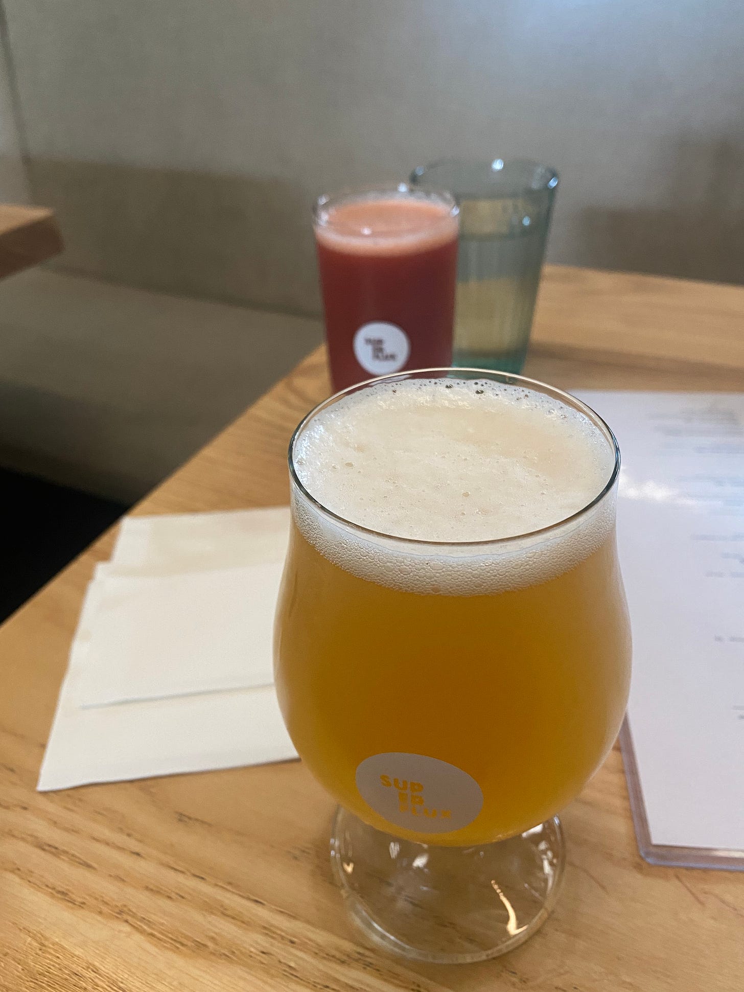 Two beers in Superflux-branded glasses, on a wooden table across from each other. The near one is an IPA in a bell-shaped glass, and the rear one is a deep red smoothie sour in a half-pint glass. A menu in a plastic sleeve is visible on the right-hand side of the table.