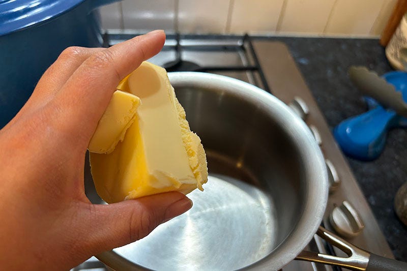 A hand holding a stick of butter, about to place it in a saucepan.