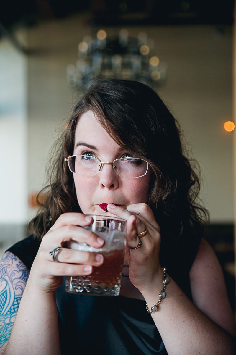 Picture of Caitlin Starling. She has wavy hair, glasses, and a brightly colored arm tattoo. She is sitting in a room with a blurry chandelier in the background and sipping a drink.