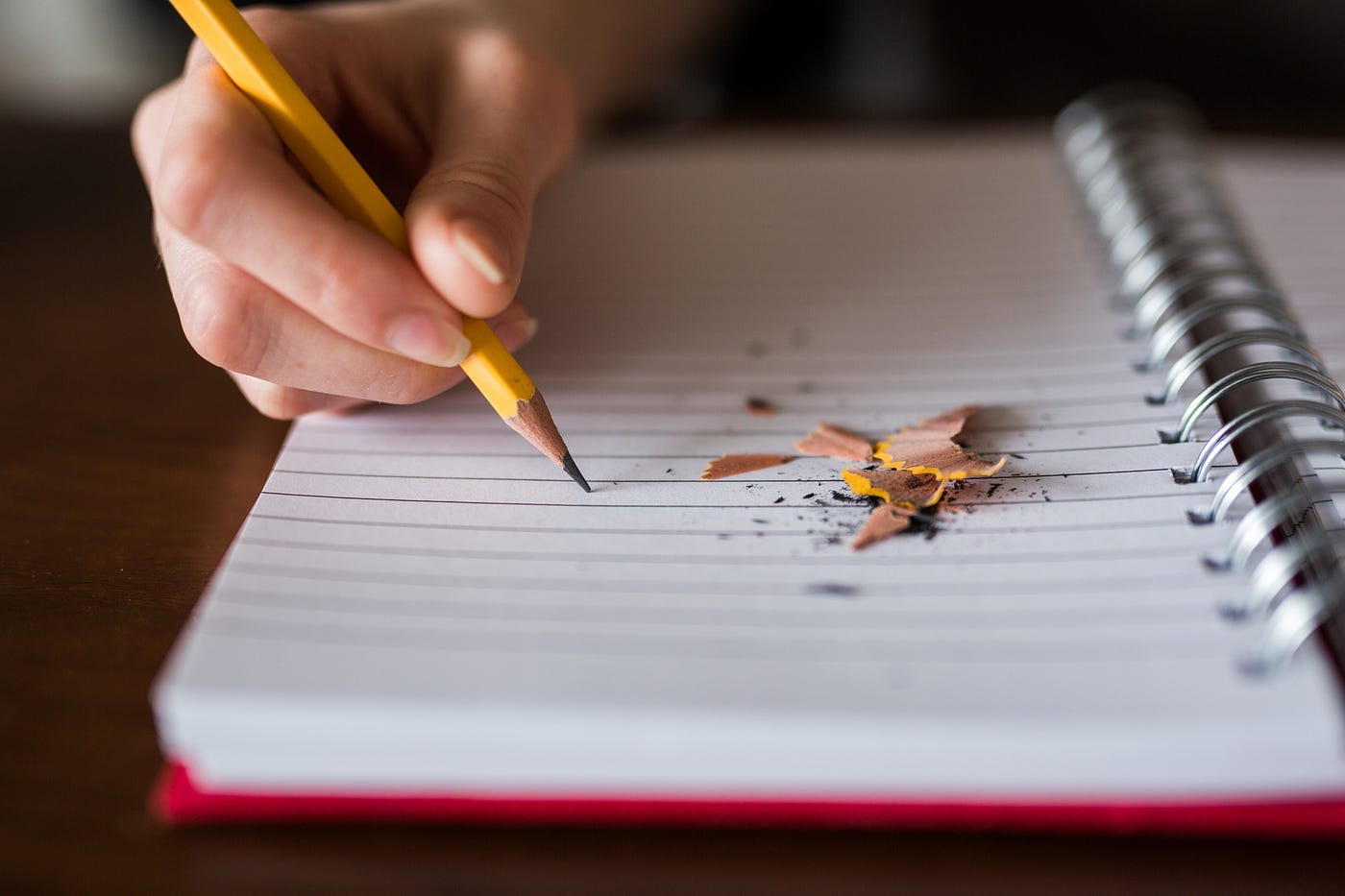 A writer is writing with a yellow pencil in a lined spiral bound notebook, lying on the notebook are pencil shavings.
