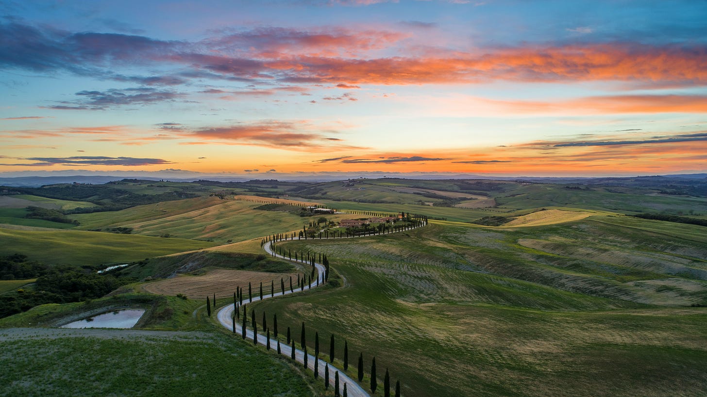 A view of a sunset in Tuscany showing a wide and wondrous landscape. A tree-lined road winds its way across this landscape. There are rolling green hills, valleys, a lake, a glimpse of a river, in the distance a building with windows, and far beyond all these, cloud-shrouded mountains and the sky streaked with clouds and the orange of the setting sun.