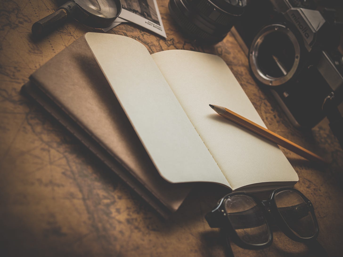 Image is a photo of an open notebook with blank pages atop a folder and a map, surrounded by various objects like a magnifying glass, an old manual camera, a pencil, and glasses.