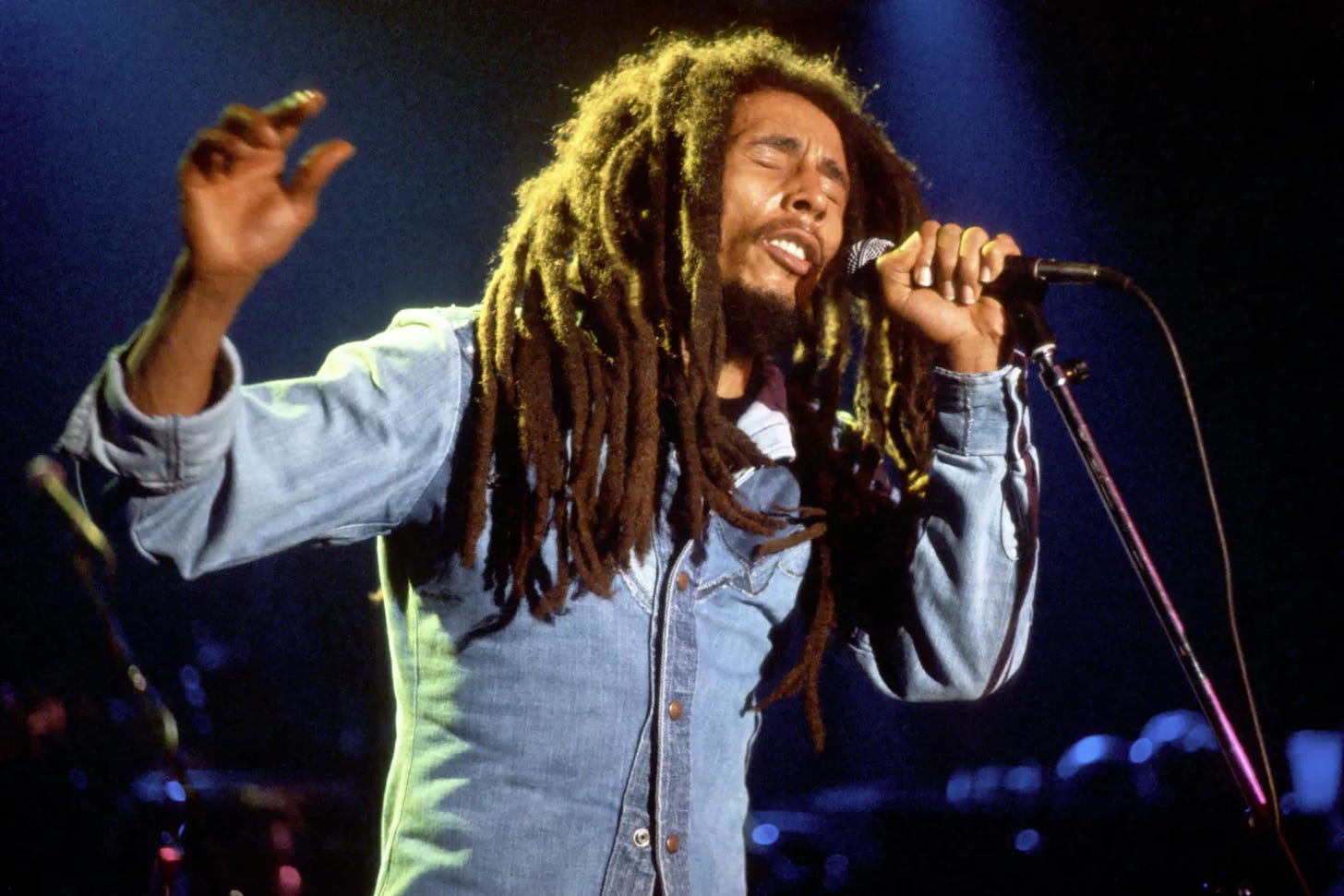 A picture of Bob Marley holding a mic with his left hand and raising his right hand up while passionately singing to an audience not visible in the image