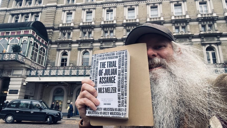 A white man with a long fuzzy white beard wears a cap and holds up a copy of the Trial of Julian Assange by Nils Melzer.