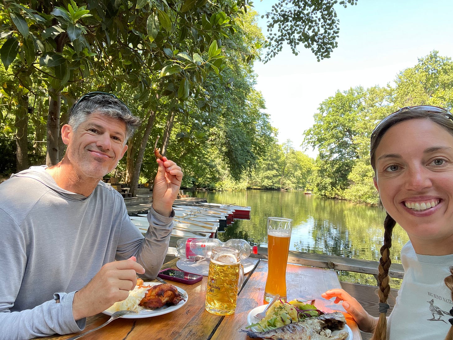Grice and Mina eating German fare with beers at a wooden table overlooking a lake with boats.