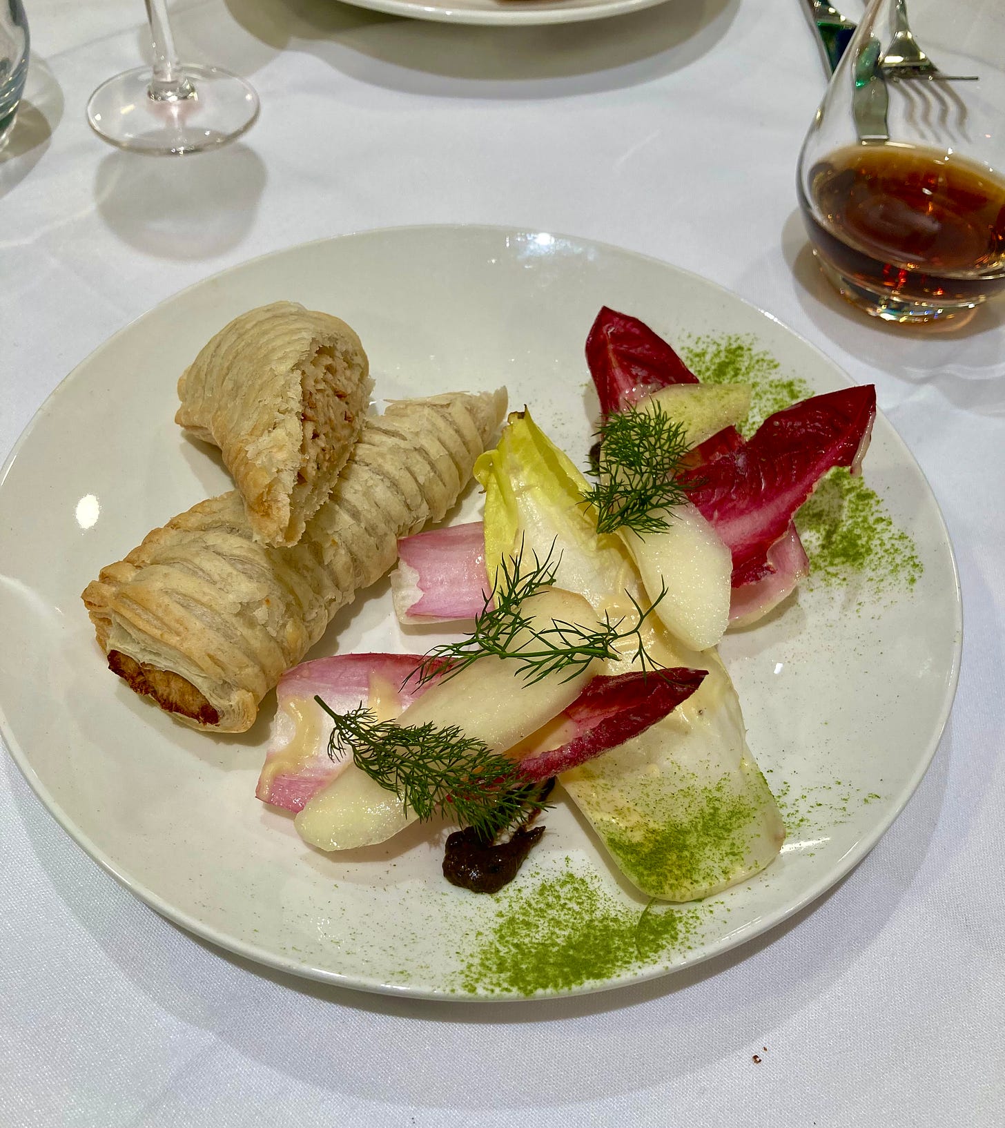 photograph of a vegan sausage role, cut diagonally and beautifully placed on a plate next to a colourful chicory and pear salad dusted with green powder and sitting atop an atful smear of picked ketchup