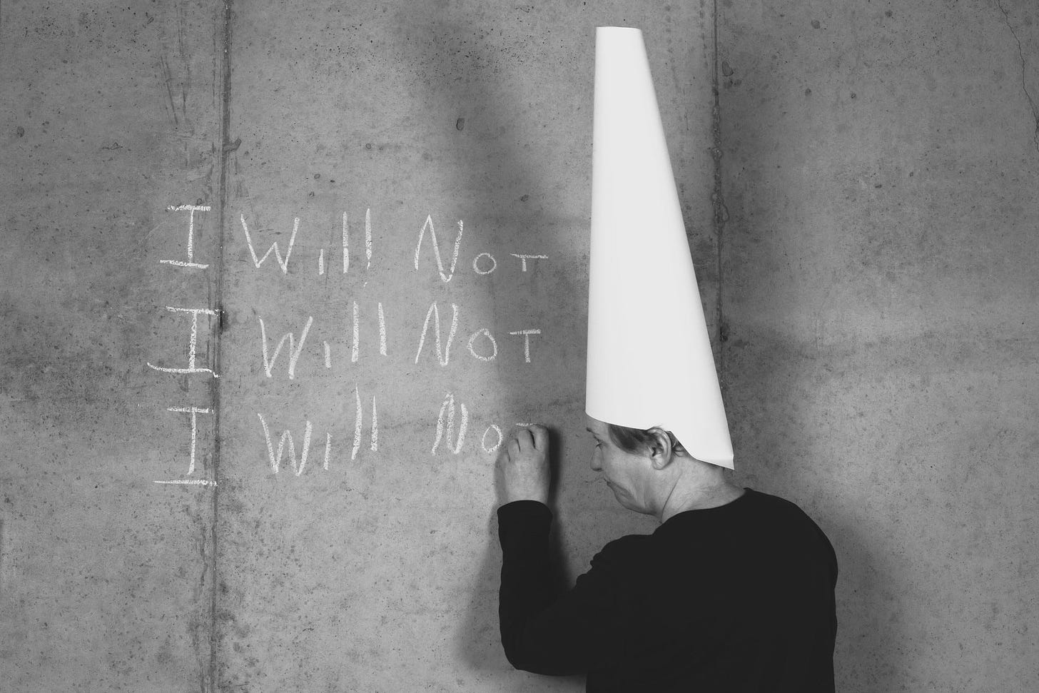 Man in a dunce cap standing before a chalkboard on which he has written, “I Will Not” three times.