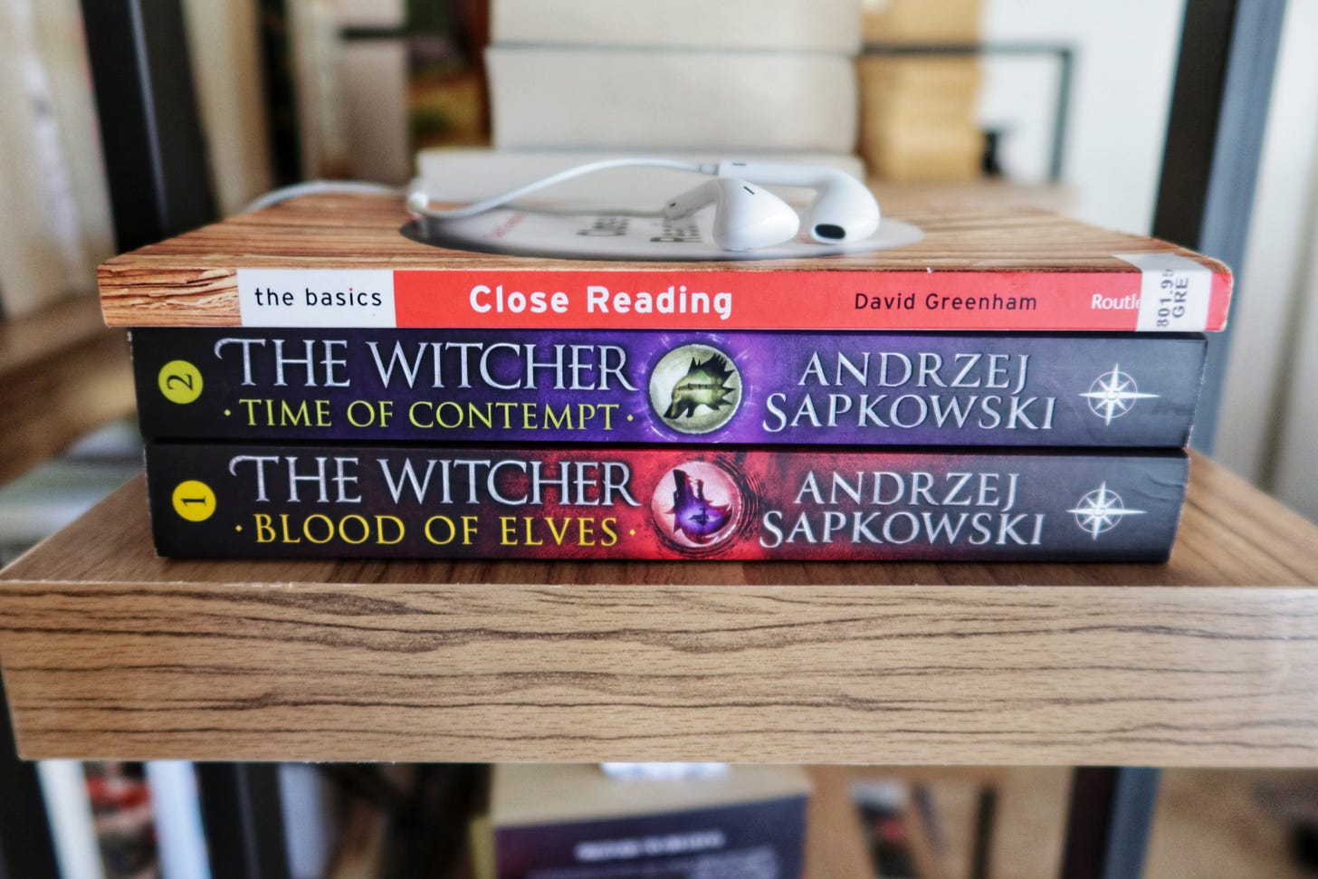 Small pile of books with earbuds on top. Books (top to bottom): CLOSE READING: THE BASICS by David Greenham; THE WITCHER TIME OF CONTEMPT by Andrzej Sapkowski; THE WITCHER BLOOD OF ELVES by Andrzej Sapkowski.