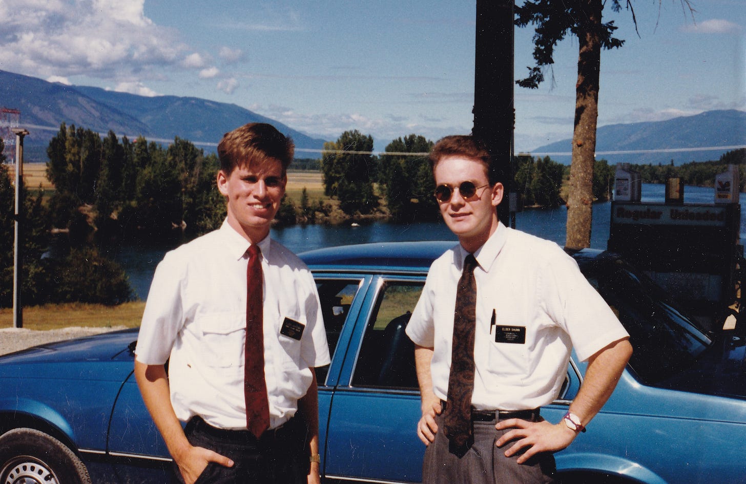 Two young male Mormon missionaries pose in front of a blue Chevy Cavalier against a backdrop of water, trees, fields, mountains, and cloudy blue sky.