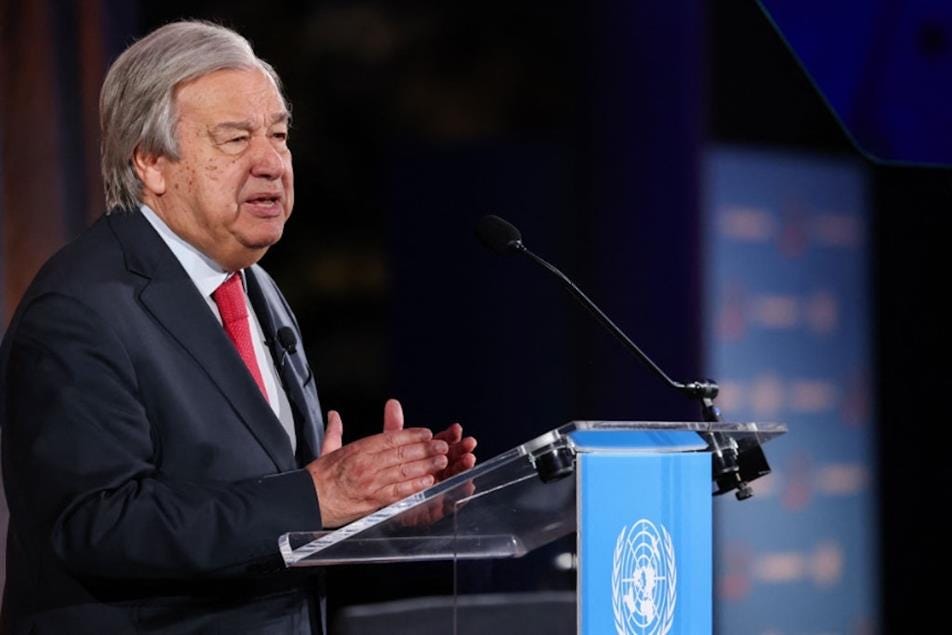 UN Secretary General António Guterres, in a dark suit and red tie, gestures from behind a lectern adorned with the blue UN flag.