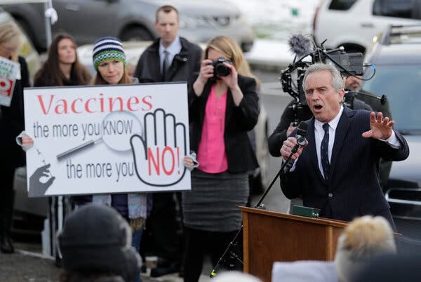 Robert F. Kennedy Jr. holds a microphone in one hand and gestures with the other while speaking outdoors behind a wooden lectern. A person in the crowd next to him carries an anti-vaccination sign.