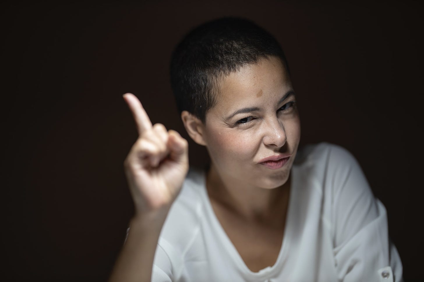 A woman with very short hair, in a white shirt, is giving the finger toward the camera.