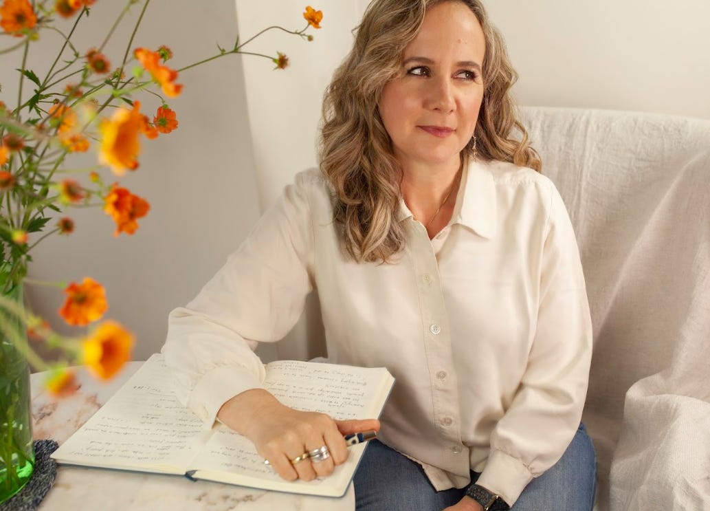 A middle-aged woman with curly, long, blonde hair is sitting on a sofa and her arm is placed on a coffee table next to her. There's a notebook on the table, and a vase with orange flowers on one side.