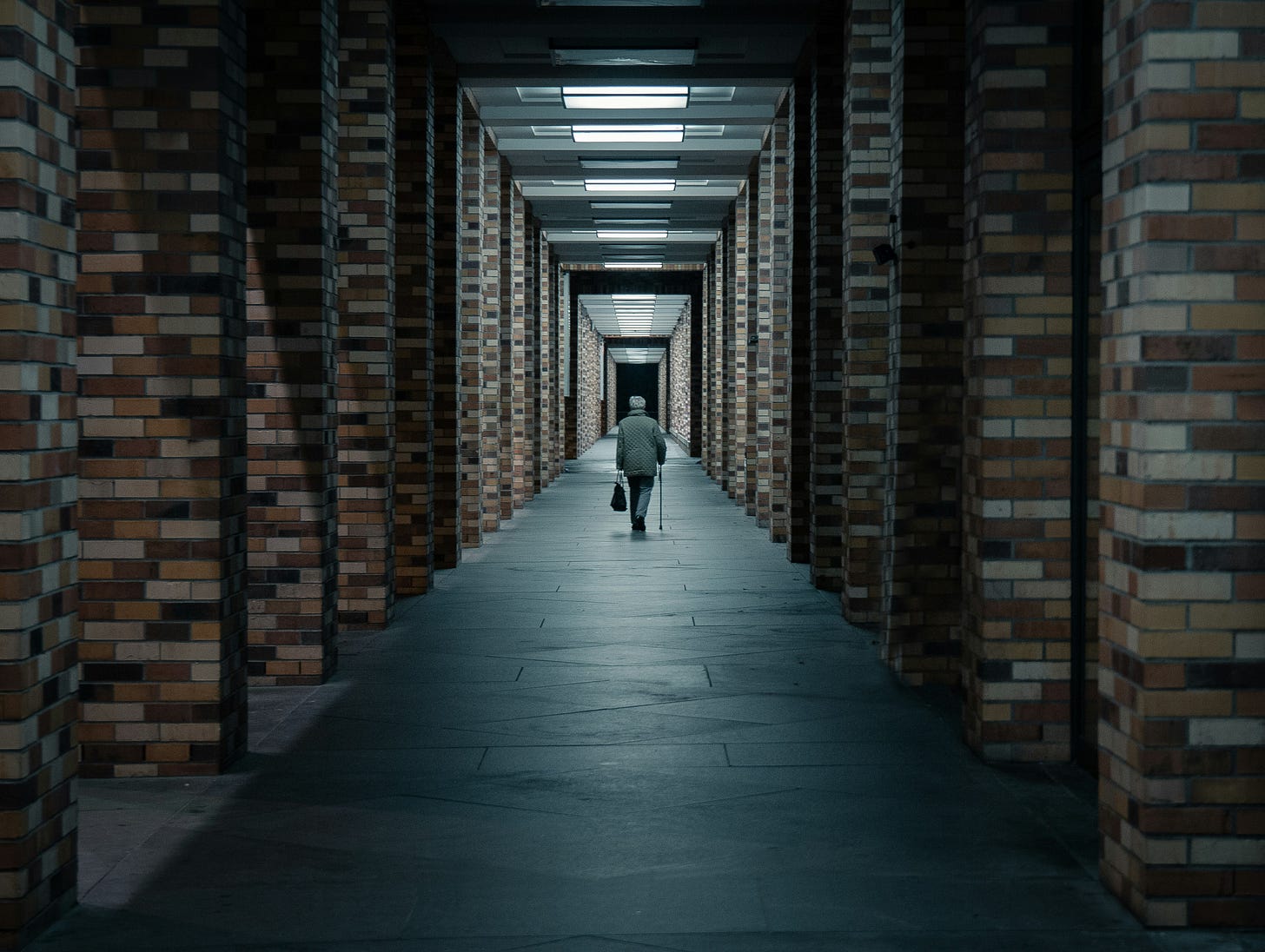 A man with a cane walks down a long hallway lined with brick pillars. 
