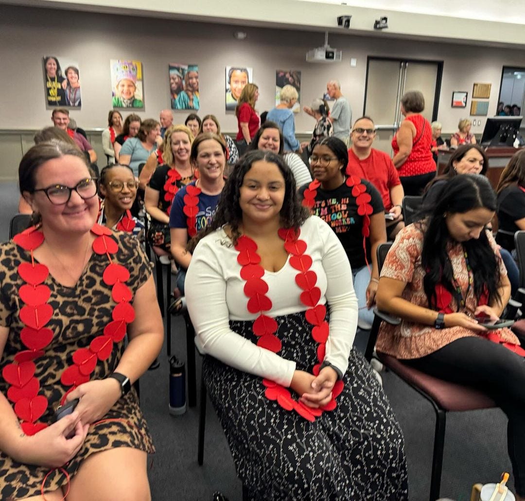 Large group of smiling people sitting in the audience of the PUSD Board Meeting. They are wearing necklaces made of large red hearts around their necks to symbolize the 1800 students that lost services. 