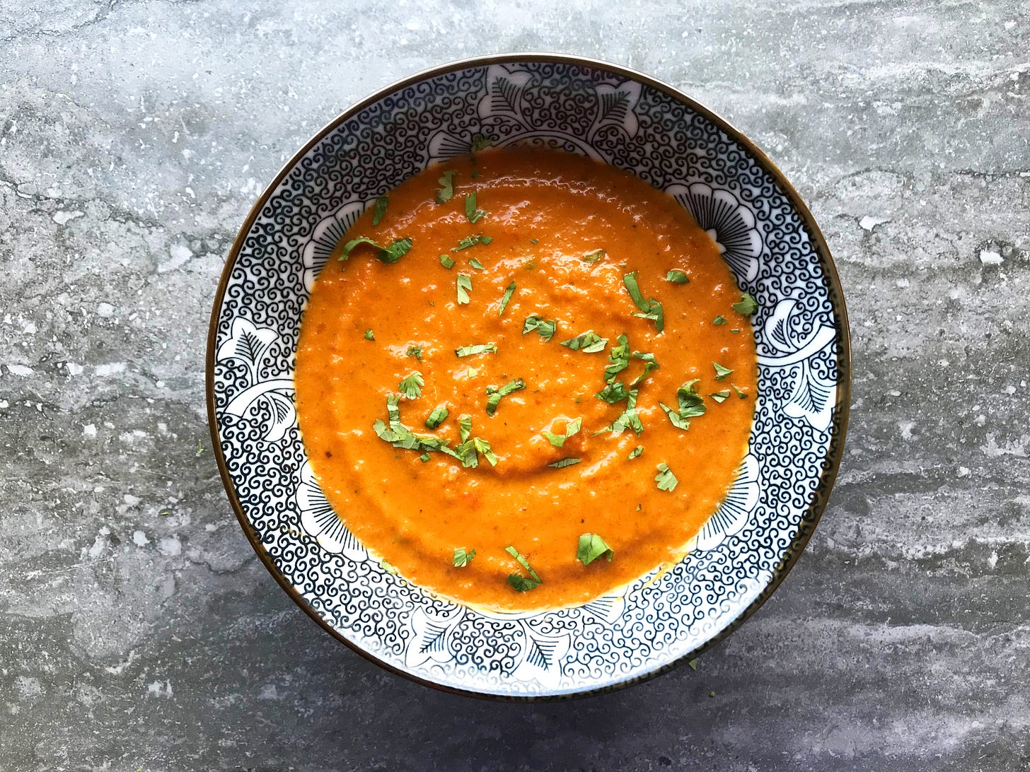 A bowl of Roasted Honey-Harissa Carrot Soup, flecked with minced coriander. The bowl's inside is a black line drawing in a floral pattern, which contrasts against the bright reddish orange of the soup.