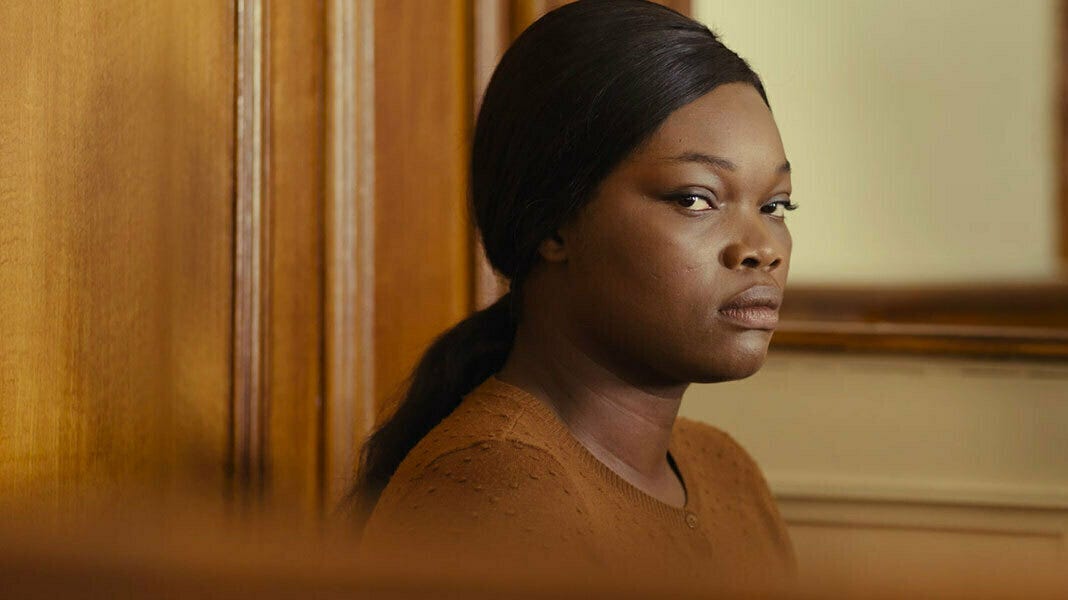 A Black woman with a low ponytail dressed in a brown cardigan sits in a courtroom defendant box and gazes towards the camera. Her skin, sweater and the wood of the courtroom are all varying but similar shades of brown.