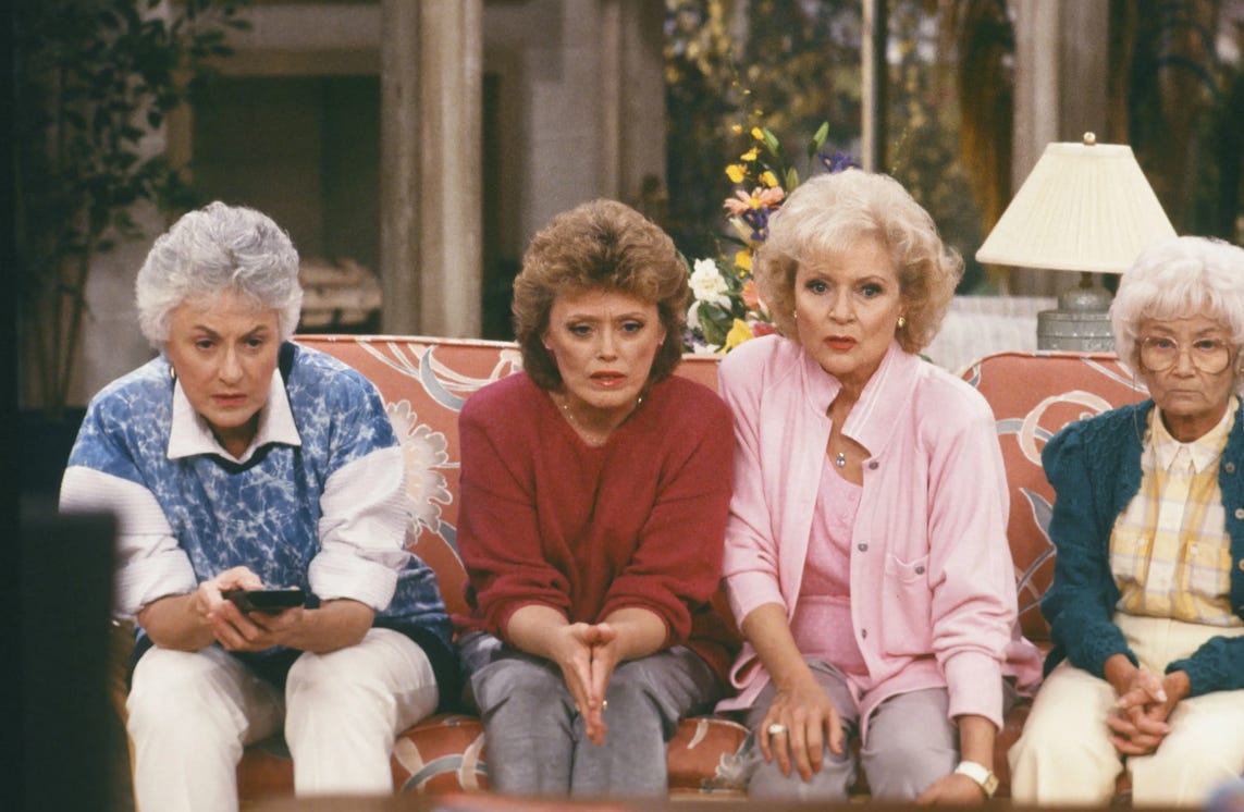 the cast of The Golden Girls sitting on a couch, seeming to watch a TV that is below the frame of view. L to R: Dorothy, Blanche, Rose, Sophia