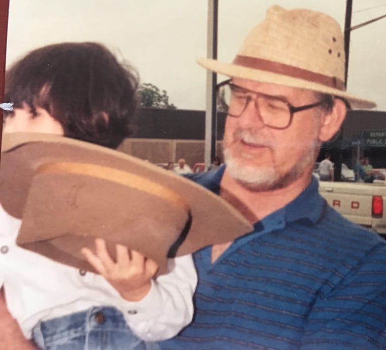 Photo of Lyric, dressed in jeans and a white shirt, holding a big brown hat, as their grandfather carries them, while wearing his own hat.