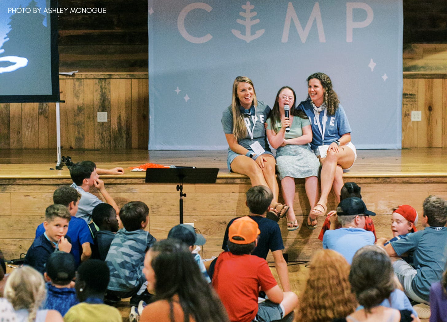 Penny sits on stage between two young adults. She holds a mic and is singing while a group of kids sits in front of her listening.