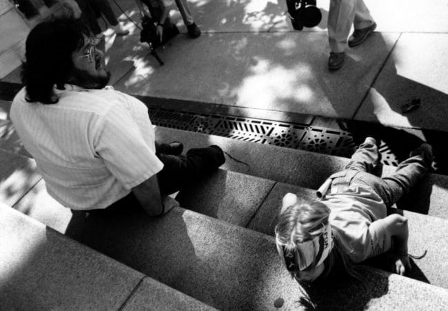 In a black and white photo, two disabled people are beginning to climb stairs. One white man is moving backwards and a young white girl is on her stomach.